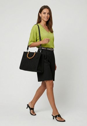 Nainen kävelee, päällään limenvihreä V-kauluksinen paita, mustat shortsit, mustat korolliset sandaalit ja kantaa mustaa bambukahvallista käsilaukkua olallaan.