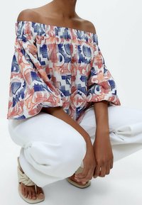 Off-the-shoulder patterned blouse in white, red, and blue. Features puff sleeves and a relaxed fit, paired with white pants and beige sandals.