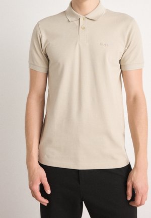 Mann trägt beige kurzärmeliges Poloshirt mit Kragen und schwarze Hose, steht vor einfachem hellem Hintergrund.