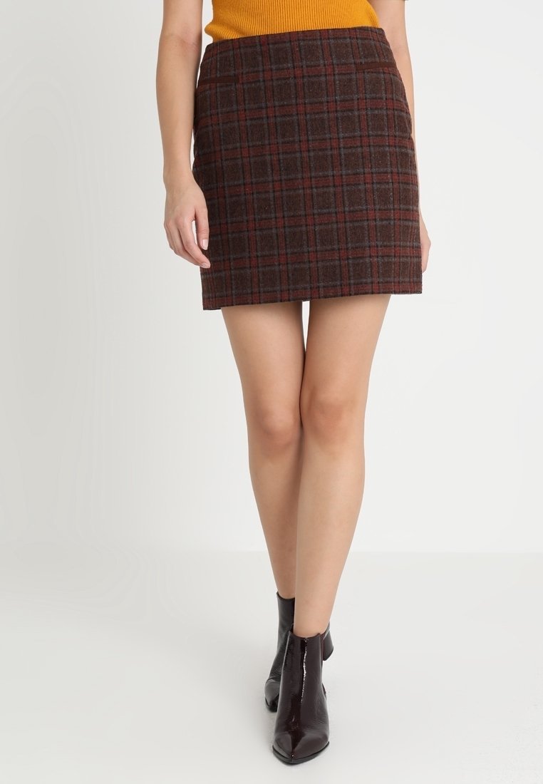 Femme portant une jupe courte à carreaux marron, un haut moutarde et des bottines marron foncé, debout sur un fond clair uni.