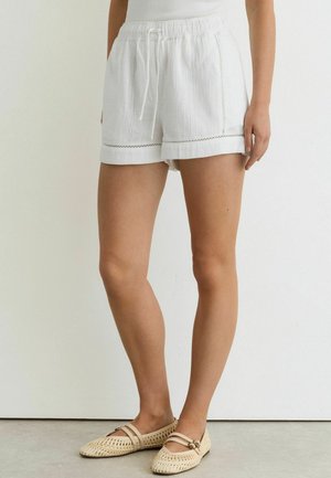 Kvinde iført hvide snøre-shorts med dekorative syninger og beige vævede flade sko, stående på en grå gulv mod en hvid væg.