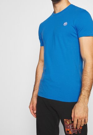Homme portant une chemise bleue ajustée à manches courtes et un pantalon noir avec le texte orange "SUPERDRY" sur la cuisse gauche, debout devant un fond uni.