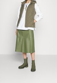 Oliwkowa pikowana kamizelka z kapturem na białej bluzie, zestawiona z pasującą zieloną skórzaną midi spódnicą i botkami za kostkę.