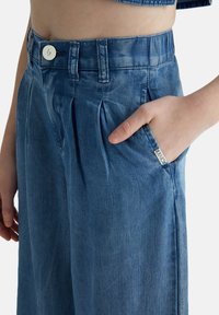 Pantaloni in denim azzurro chiaro con fronte plissettato, vita elasticizzata, tasca laterale e dettaglio della etichetta con logo. Texture liscia e design casual.