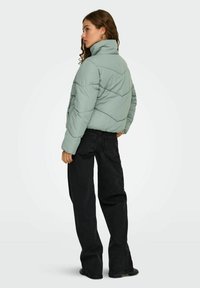Grøn pufferjakke med høj krave og quiltet design, parret med sorte brede bukser. Har struktureret stof og neutrale farver.