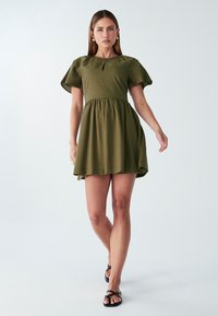 Olive zeleno kratka obleka s gathered krilom, dekoltejem v obliki ključa in mehko, tkano teksturo. V kombinaciji z črnimi zavezankami.