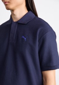 Námornícka polo košeľa vyrobená z textúrovanej látky s plochým golierom a krátkymi rukávmi. Obsahuje malé fialové logo Puma na hrudi.