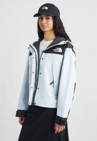 Ανοιχτό μπλε μπουφάν με μαύρες λεπτομέρειες, κλείσιμο με φερμουάρ και λογότυπο στο στήθος. Φορεμένο πάνω από λευκό πουκάμισο, σε συνδυασμό με μαύρο καπέλο. Λείο ύφασμα.