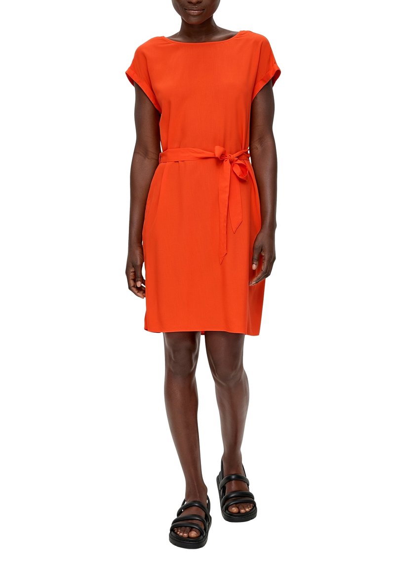 Oranje jurk met korte mouwen, een aangesloten taille en een strik aan de voorkant, gemaakt van een gladde, lichtgewicht stof. Zwarte sandalen worden apart gedragen.