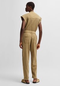 Khaki jumpsuit with short sleeves, cinched waist, and two back pockets. Soft, textured fabric with a relaxed fit.