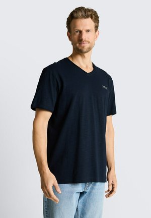 Mężczyzna z krótkimi kręconymi włosami, ubrany w czarną koszulkę z dekoltem w serek i jasnoniebieskie dżinsy, stojący na jednolitym jasnoszarym tle.