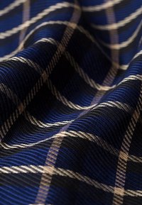 Zbliżenie na niebieską, czarną i beżową tkaninę w kratę z prążkowaną teksturą i widocznymi fałdami tworzącymi światła i cienie.