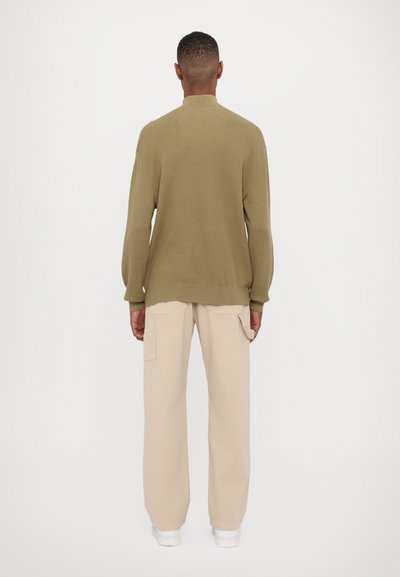 Knit olive-green sweater with a high collar, long sleeves and tapered cuffs, paired with beige cargo pants and white sneakers.