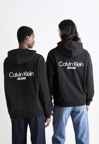 Schwarze Kapuzensweatshirts mit dem weißen "Calvin Klein JEANS"-Logo auf dem Rücken. Getragen mit locker sitzenden blauen Jeans, die einen lässigen Stil zur Schau stellen.