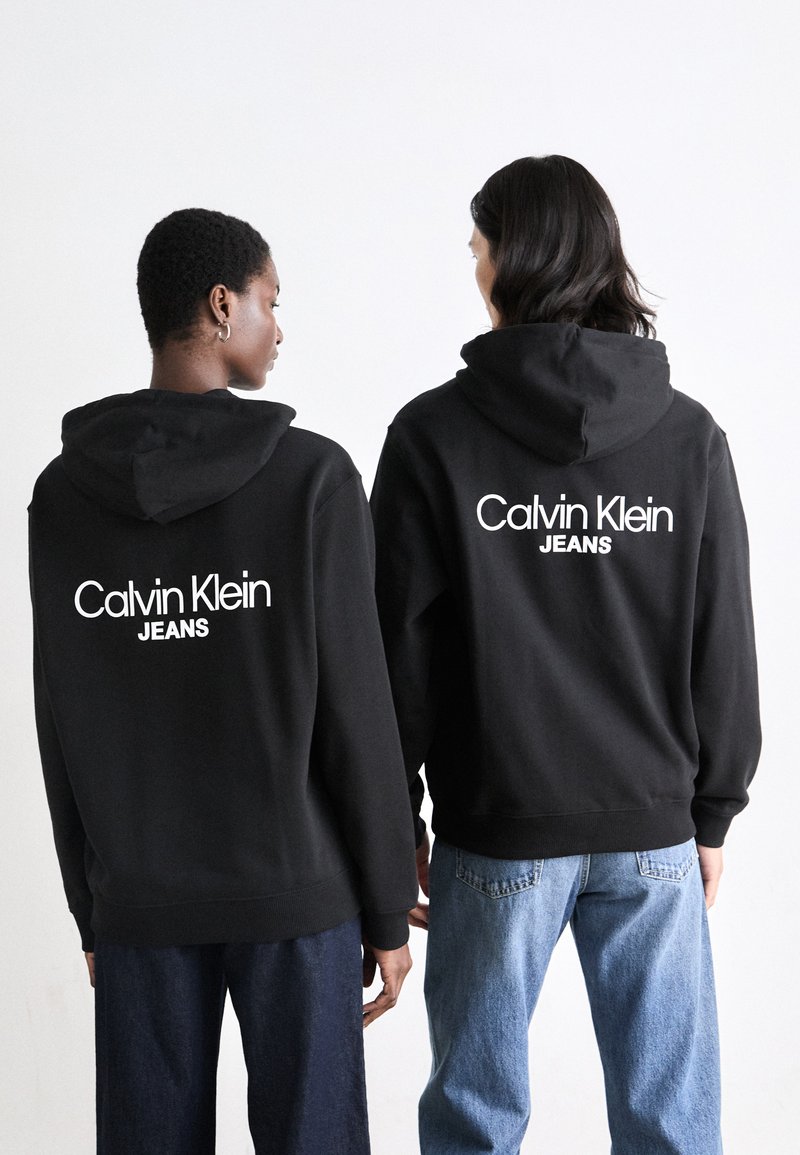 Schwarze Kapuzensweatshirts mit dem weißen "Calvin Klein JEANS"-Logo auf dem Rücken. Getragen mit locker sitzenden blauen Jeans, die einen lässigen Stil zur Schau stellen.