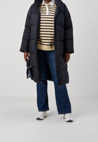Eine Person trägt einen langen schwarzen Puffer-Mantel über einem beigefarbenen gestreiften Pullover, blauen Jeans, weißen Sneakers und hält eine kleine marineblaue Handtasche.