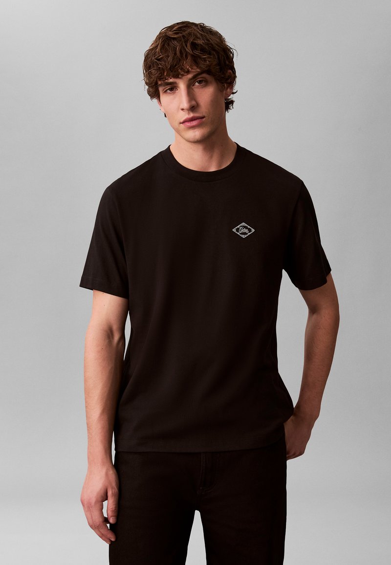 T-shirt en coton noir avec col rond, manches courtes et petit logo en forme de losange sur le côté gauche de la poitrine. Texture lisse et coupe ajustée.
