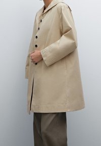 Manteau beige léger avec un grand col, une fermeture à bouton à l'avant et des fentes sur les côtés. Conçu avec une coupe décontractée et des manches longues.