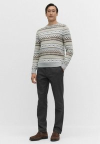 Man staat met handen in zijn zakken, draagt een geprinte lichtblauwe, bruine en witte trui, donkere grijze broek enbruine schoenen.