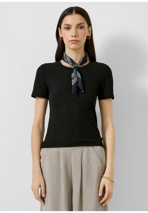 Vrouw draagt een zwart textuur kort mouw shirt, lichtbeige hoge taille broek, zwarte patroonige nek sjaal, gouden oorringen en een gouden armband.