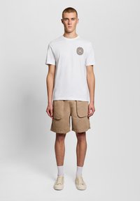 Νεαρός άνδρας που στέκεται, φορώντας λευκό κοντομάνικο μπλουζάκι με κυκλικό λογότυπο στο στήθος, μπεζ cargo σορτς, λευκές κάλτσες και μπεζ αθλητικά παπούτσια.