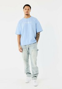 Camiseta de gran tamaño en azul claro con texto bordado, combinada con pantalones de mezclilla deslavados y zapatillas blancas. Diseño de pierna recta y ajuste casual.