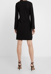 Czarna sukienka z długim rękawem o dopasowanej sylwetce, z okrągłym dekoltem i łańcuszkowym akcentem z tyłu, wykonana z gładkiego materiału.