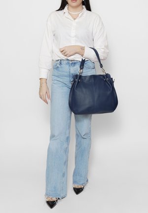 Femme portant un chemisier blanc et un jean bleu clair tenant un grand sac à main en cuir bleu marine par ses poignées.