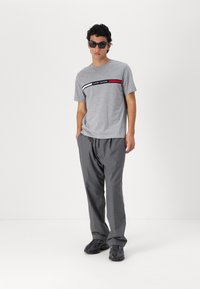 Grå t-shirt med et horisontalt logo-bånd i rød, hvid og sort, parret med løse grå bukser og sorte teksturerede sneakers.