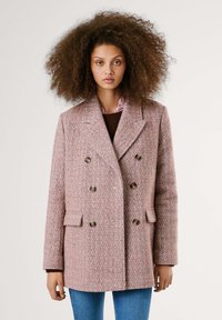 Manteau croisé en motif chevron rose, avec de grands boutons marron, des revers ombragés et deux poches avant. Porté par-dessus un pull marron.