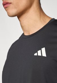 Musta urheilupaita, jossa on pyöreä pääntie ja valkoinen kolmiraitainen logo vasemmassa rinnassa. Pehmeä ja sileä kangaspinta.