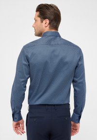 Camicetta blu con un piccolo motivo geometrico, vestibilità aderente, maniche lunghe e colletto classico, abbinata a pantaloni scuri color navy.