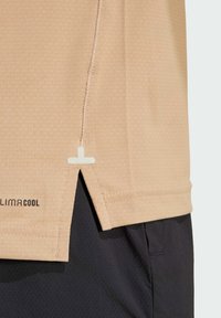 Tan athletic shirt with a textured fabric, short sleeves, side slits, and a white accent. Features a logo at the bottom left.