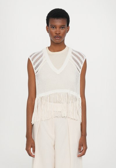 Top de punto sin mangas en color crema con escote en V, que presenta patrones de punto abierto y un dobladillo con flecos. Se lleva sobre una camisa clara y se combina con pantalones en tonos claros.