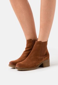 Brown suede ankle boots with elastic side panels and a textured block heel. Smooth surface with rounded toe, designed for easy wear.