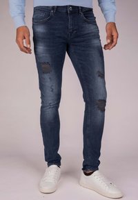 Dark blue slim-fit jeans with distressing and patches on the legs, featuring a classic five-pocket design and a zip fly closure.