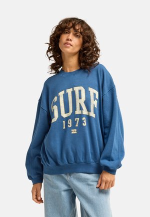 Frau mit lockigem Haar, die einen blauen Pullover mit dem Text "SURF 1973" und hellblaue Jeans trägt, steht vor einem einfarbigen Hintergrund.
