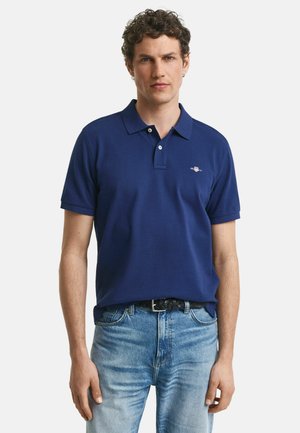 Navyblaues Polo-Shirt aus Baumwolle mit einer Zwei-Knopf-Leiste, kurzen Ärmeln und einem kleinen Logo auf der linken Brust. Getragen mit blauen Jeans.