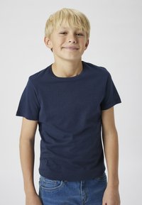 Náměsně modré bavlněné tričko s krátkým rukávem a kulatým výstřihem, nošené s světle modrými džíny, umístěné na světlém pozadí.