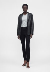 Große Frau trägt schwarzen Lederblazer, hellgraues T-Shirt, schwarze Skinny-Hosen und schwarze Sandaletten mit hohen Absätzen, steht vor einem schlichten weißen Hintergrund.