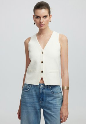 Witte mouwloze vest met een V-hals, met een knoopsluiting aan de voorkant en twee zijzakken. Draag het over lichtblauwe spijkerbroek. Gladde textuur.