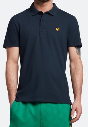 Mężczyzna w ciemnogranatowej koszulce polo z krótkim rękawem z żółtym logo ptaka oraz jasnych zielonych spodenkach w kratę, stojący z rękami w kieszeniach.