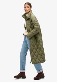 Manteau long matelassé vert olive avec col montant ; porté avec un pull crème et un jean bleu clair ; associé à des bottes bordeaux à lacets.