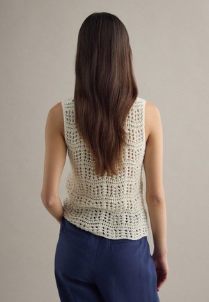 Woman with long brown hair wearing a sleeveless cream crochet top and dark blue pants, standing against a plain beige background.
