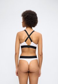 Γυναίκα με σγουρά μαλλιά που φοράει λευκό και μαύρο σουτιέν με λουράκια σε σχήμα σταυρού και ταιριαστά εσώρουχα με ψηλή μέση, γυρισμένη προς τα πίσω μπροστά σε απλό φόντο.