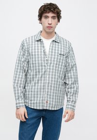 Καρό πουκάμισο με μακριά μανίκια σε αποχρώσεις του μπλε και λευκού, βαμβακερό ύφασμα, κουμπιά μπροστά και κεντημένο λογότυπο στην τσέπη του στήθους. Φορεμένο με μπλε τζιν.