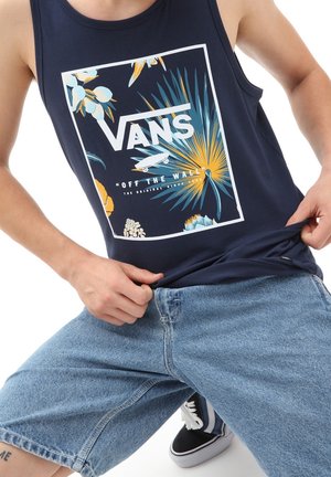 Personne portant un débardeur bleu marine avec un imprimé floral du logo Vans et un short en jean bleu, tirant légèrement le débardeur vers l’extérieur.