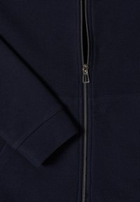 Primo piano di un tessuto lavorato a maglia blu navy con zip in metallo argentato e dettaglio di polsino a manica lunga.