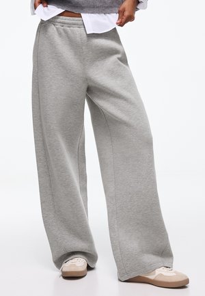 Personne portant un pantalon de survêtement gris ample et des baskets beiges, debout, une main soulevant une couche supérieure grise sur une chemise blanche.