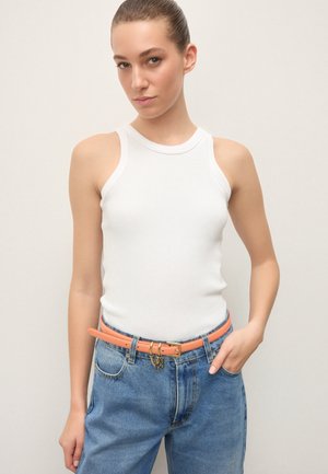 Joven con el cabello peinado hacia atrás, vestida con una blusa blanca sin mangas, jeans azules de talle alto y un cinturón coral con un llavero colgante.